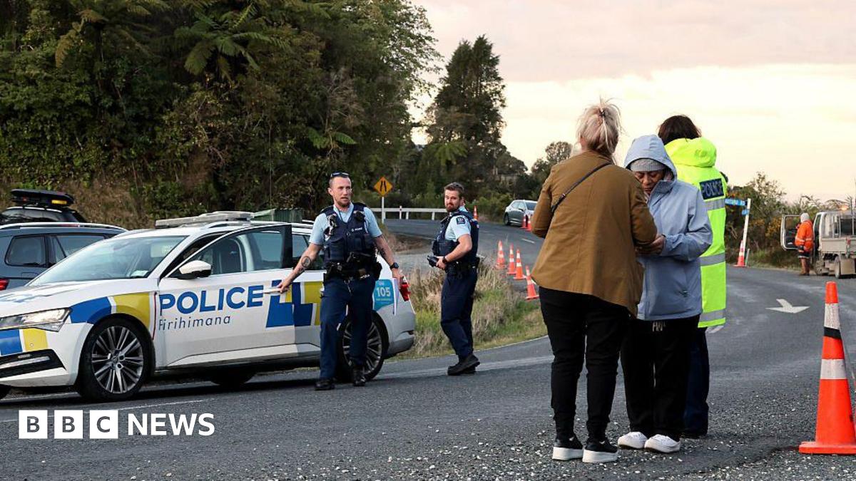 Two policemen standing near a police car, while two women wearing jackets and long pants stand on the side of the road. In the background is a tall green forest.