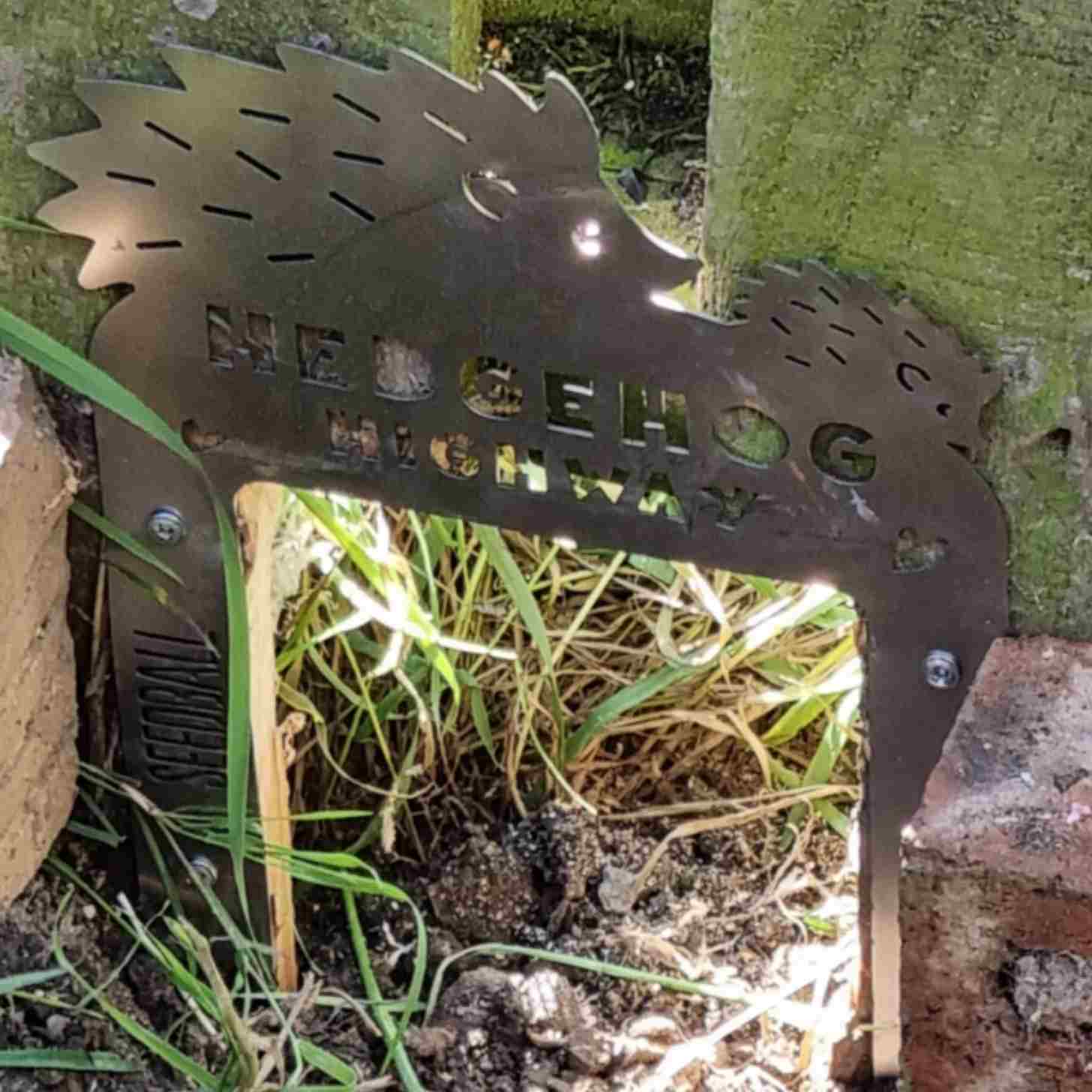 a hole in a fence with a metal sign shaped like two hedgehogs that reads: Hedgehog Highway