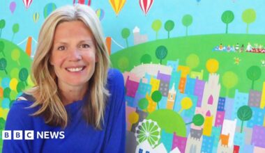 Wearing a blue top, Jenny smiles at the camera whilst posing in front of one of her artworks. She has blonde hair hanging loose below her shoulders. The art depicts hot air balloons flying over Bristol. The Clifton Suspension Bridge can be seen in the work behind her, as can children playing in a park.