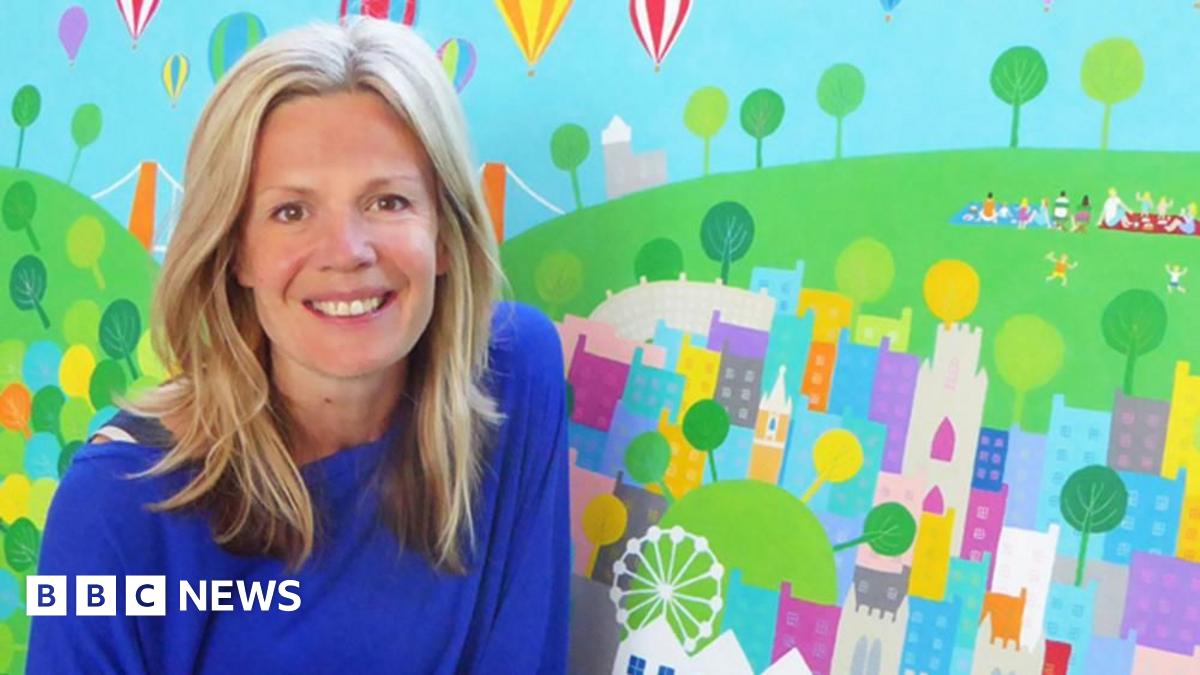 Wearing a blue top, Jenny smiles at the camera whilst posing in front of one of her artworks. She has blonde hair hanging loose below her shoulders. The art depicts hot air balloons flying over Bristol. The Clifton Suspension Bridge can be seen in the work behind her, as can children playing in a park.