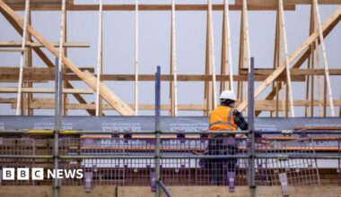 A smiling Steve Reed in suit and tie waves, wearing a red baseball cap embroidered with the words "Build Baby Build!"