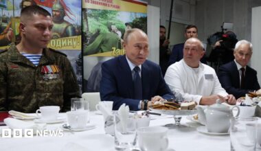Putin sitting in front of a table with tea and cakes, flanked by veterans