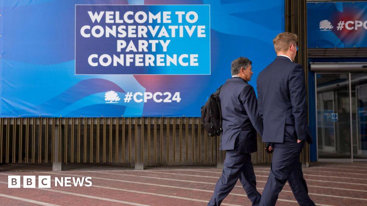 Two men in suits walk past a huge poster at the Conservative Party Conference in Manchester. The poster reads: welcome to conservative party conference.