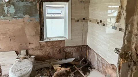Lukman Ashraf A partially demolished bathroom with exposed wooden walls showing patches of plaster, insulation, blue paint, and bare wood. Debris, including broken tiles and insulation, covers the floor. A toilet is visible in the lower left corner.