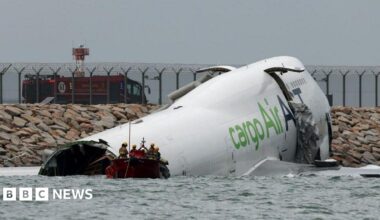 A damaged fuselage of a plane partially submerged in seawater. A small red boat carrying emergency workers is floating in front of the plane.