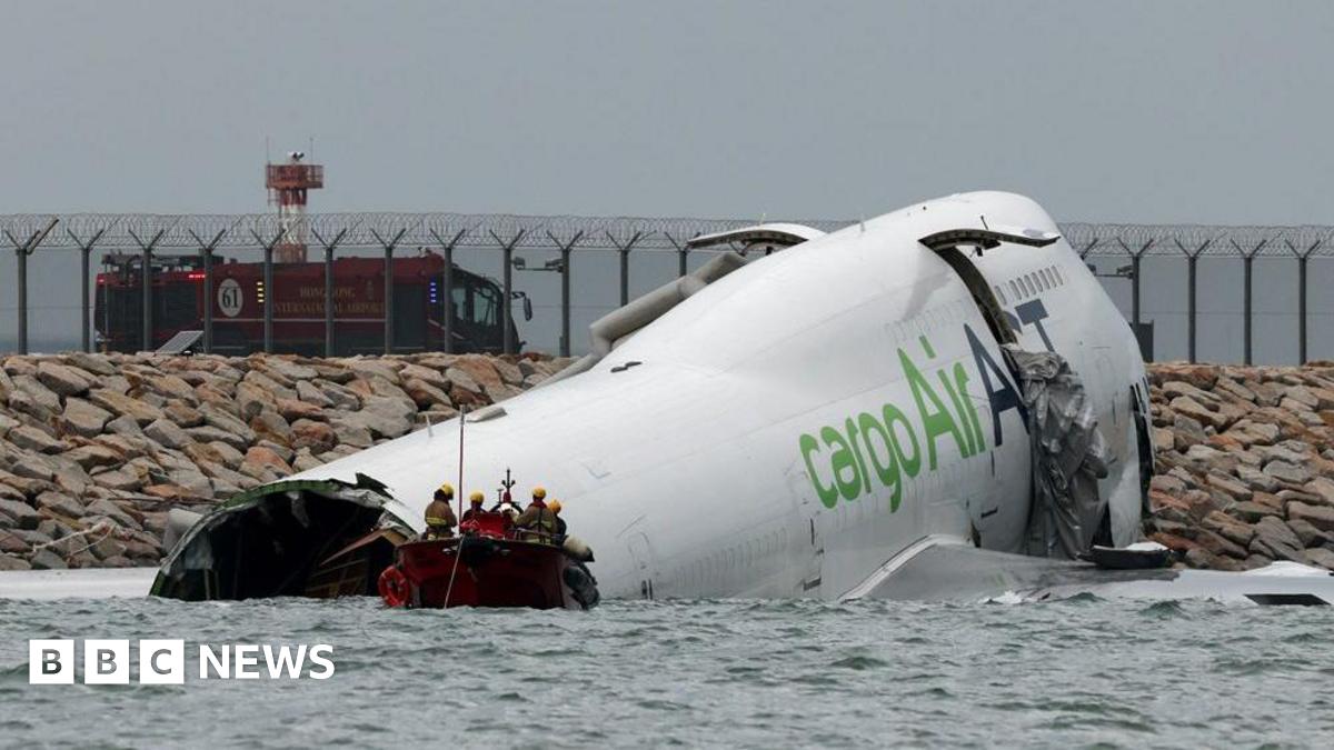 A damaged fuselage of a plane partially submerged in seawater. A small red boat carrying emergency workers is floating in front of the plane.