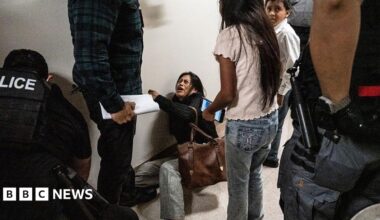 A woman lying on the floor cries. She is surrounded by officers wearing vests labelled police, and her two young children