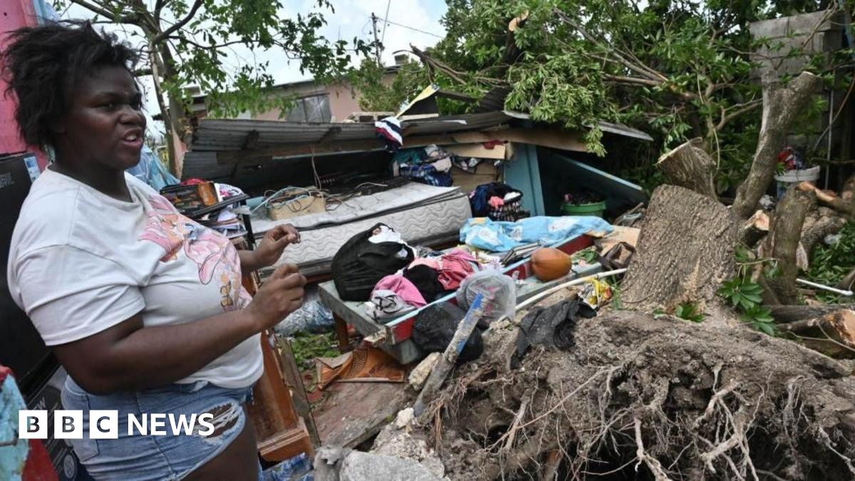 An aerial view of hurricane destruction shows collapsed buildings with roofs torn off and debris scattered across the ground. In the centre is a partially standing two-storey pink and white building. Broken wooden beams, metal sheets and concrete rubble cover the surrounding area as well as bare trees.