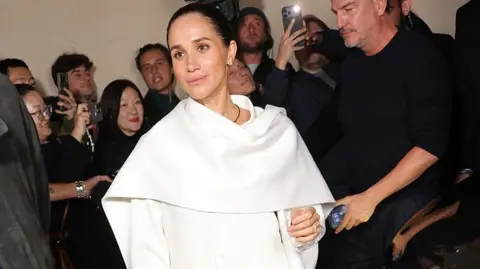 Arnold Jerocki/Getty Images for Balenciaga The Duchess of Sussex walks in front of a crowd of people dressed in black, several of whom are holding up phones. She is wearing a white jumpsuit with a cape over the shoulders. Her dark hair is tightly tied back.