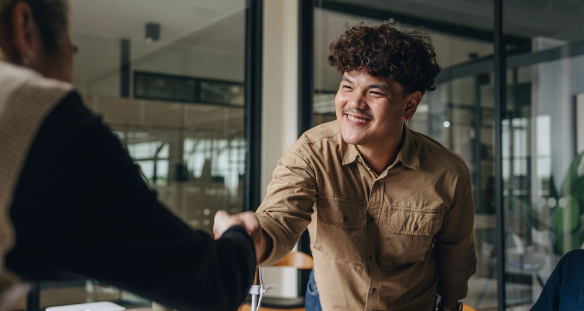 A 20-something man reaches across a table in an office to shake hands with a colleague.