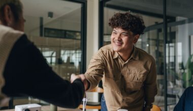 A 20-something man reaches across a table in an office to shake hands with a colleague.