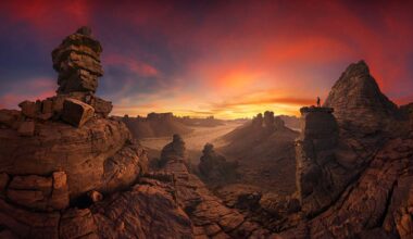 A vast desert landscape at sunset, featuring unique rock formations silhouetted against a vibrant sky with a lone figure on a peak