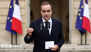 Kemi Badenoch wearing a white jacket and white t-shirt is pictured waving to the camera. There is a union jack pictured to the left.