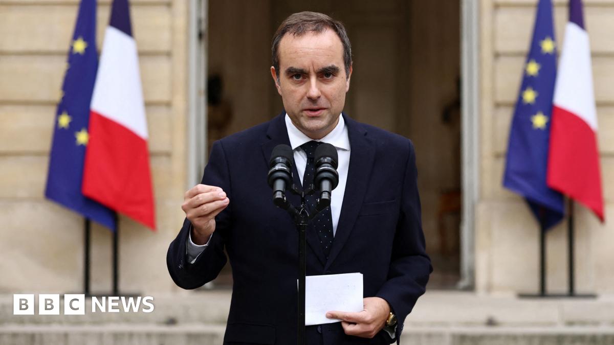 Kemi Badenoch wearing a white jacket and white t-shirt is pictured waving to the camera. There is a union jack pictured to the left.