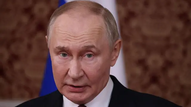 close up of Vladimir Putin in a black suit and white shirt, looking slightly to the left, Behind him, a Russian flag with the blue and white parts visible