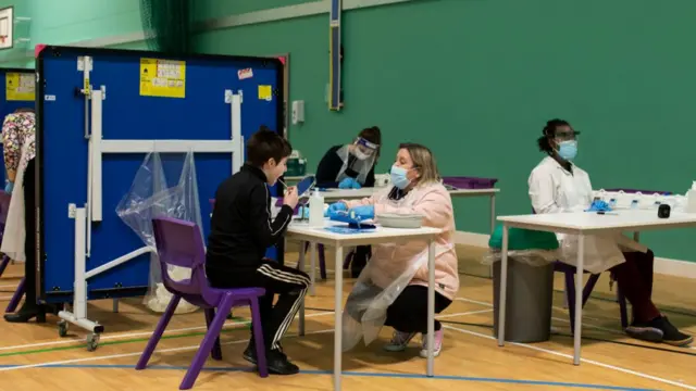 A school sports hall has been turned into a Covid testing area. There are several tables with people wearing full protective gear helping to administer tests, with a young boy in a black tracksuit completing a test.