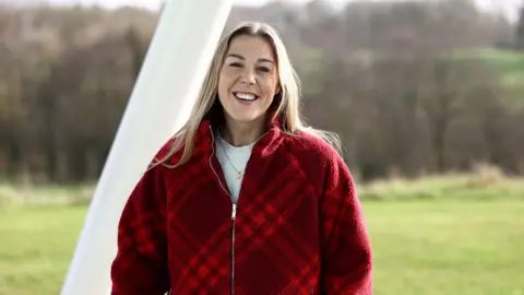 The FA via Getty Images Mary Earps smiles while looking at the camera while standing outside against a backdrop of green grass and bare trees while wearing a red zip up top over a white shirt, at St. George's Park in February 2024.
