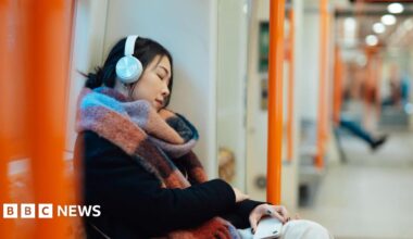 A person wearing white headphones and a large multicoloured scarf sits with her eyes closed against the window of an empty London Overground train, holding a phone in their hand.