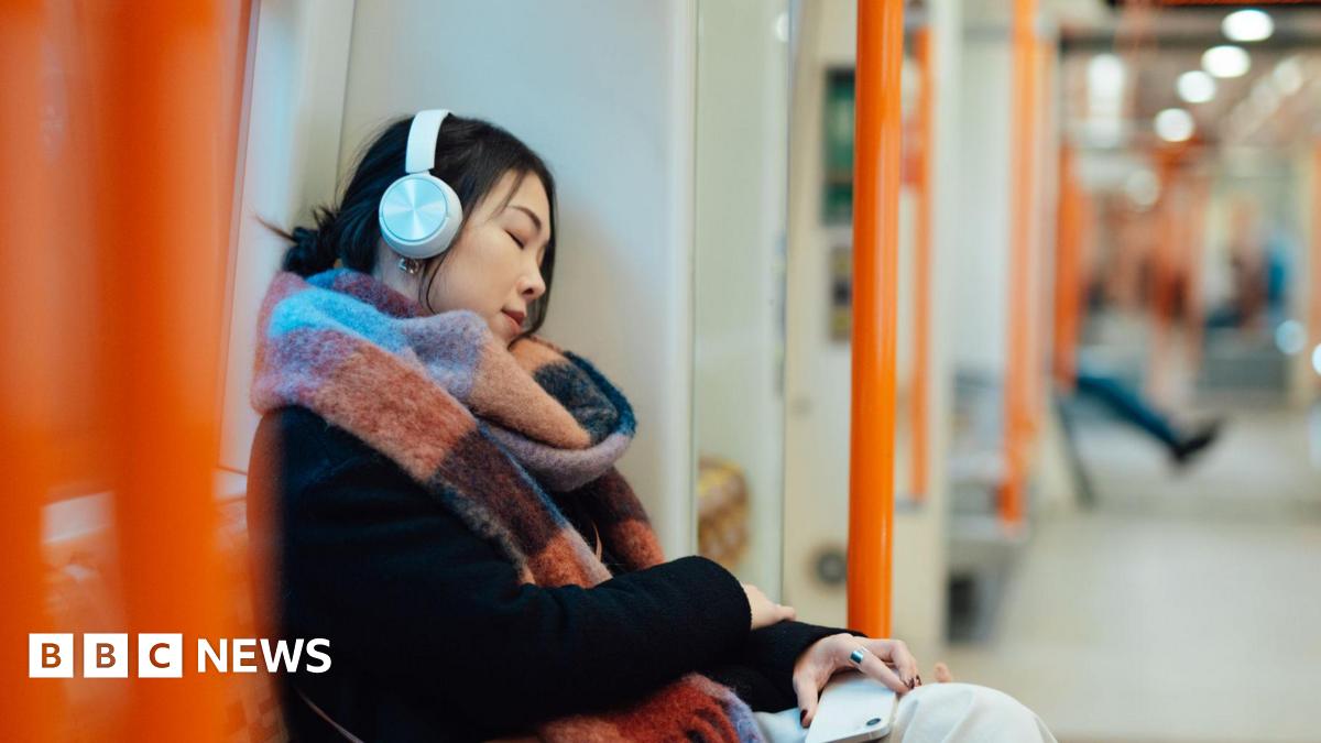 A person wearing white headphones and a large multicoloured scarf sits with her eyes closed against the window of an empty London Overground train, holding a phone in their hand.