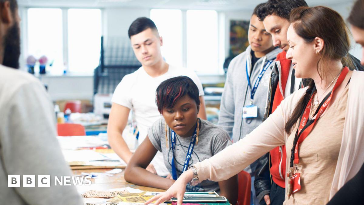 A lecturer in a classroom is pointing to a desk with different fabrics on, surrounded by a small group of students in a textiles class. They all have lanyards on their necks - the lecturer's is red and says "staff". One female student is sat at the desk close to where the lecturer is pointing, while two other male students are stood behind her.