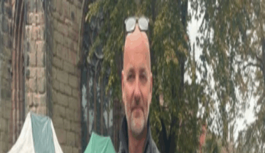 A bald man with glasses resting on his head, and wearing a black jacket and dark grey jumper, stands with his hands clasped in front of him. He is standing in front of a number of market stalls and a number of tables with bags on top of them.