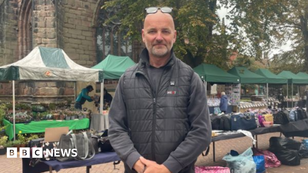 A bald man with glasses resting on his head, and wearing a black jacket and dark grey jumper, stands with his hands clasped in front of him. He is standing in front of a number of market stalls and a number of tables with bags on top of them.
