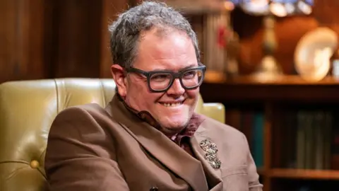 Alan Carr on the Celebrity Traitors, sitting in an armchair and smiling