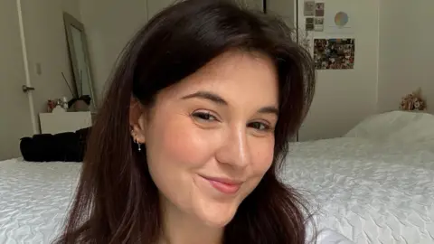 Jess Farnham Jess smiles at the camera, she has long brown hair and three earrings in her ear. She sits in front of a bed with a white duvet.
