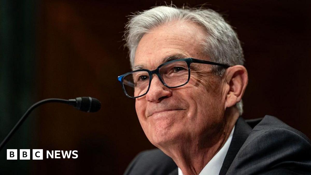 A smiling Jerome Powell, dressed in a dark grey suit, sits in front of a microphone
