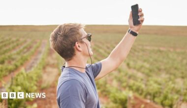 A young man in sunglasses in the countryside holds up his mobile phone to try and get better phone signal