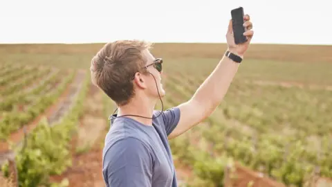 Getty Images A young man in sunglasses in the countryside holds up his mobile phone to try and get better phone signal