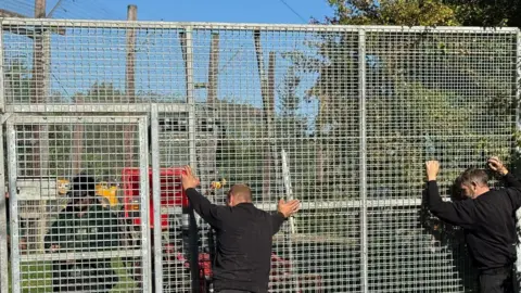Lincolnshire Wildlife Park A tall metal gate with square holes in it with two men dressed all in back leaning their hands against it.