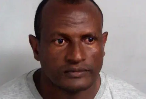 Essex Police Hadush Kebatu's mugshot. He wears a grey sweatshirt and has short black hair. He is positioned in front of a plain grey wall.