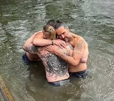 Two men embrace in a river, one with extensive back tattoos.