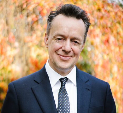 Mark Hallett smiling, wearing a dark suit, white shirt, and patterned tie, with blurred orange and red foliage in the background.