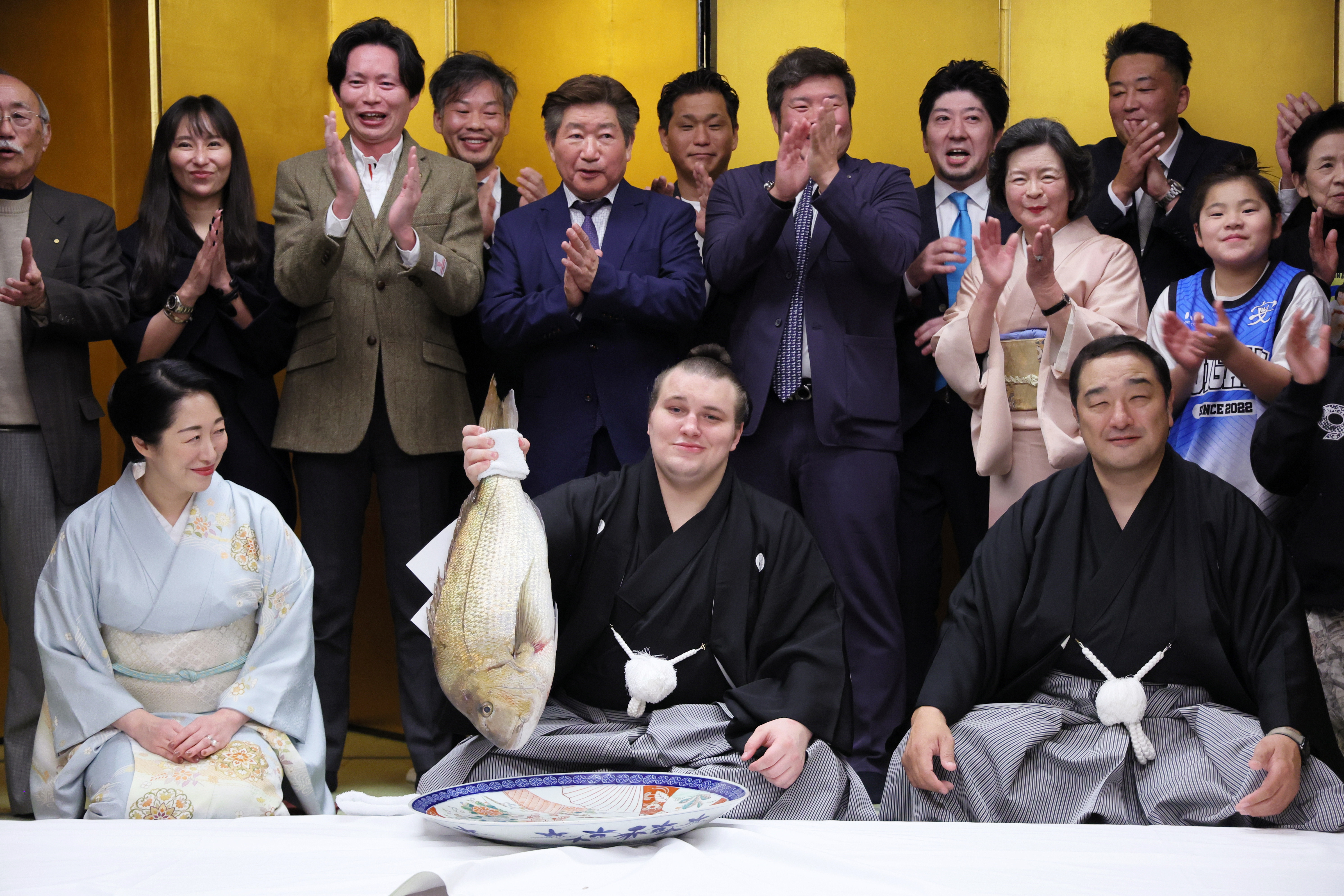 Ukrainian sumo wrestler Aonishiki holding a seabream after winning the Kyushu Grand Sumo Tournament, surrounded by people applauding.