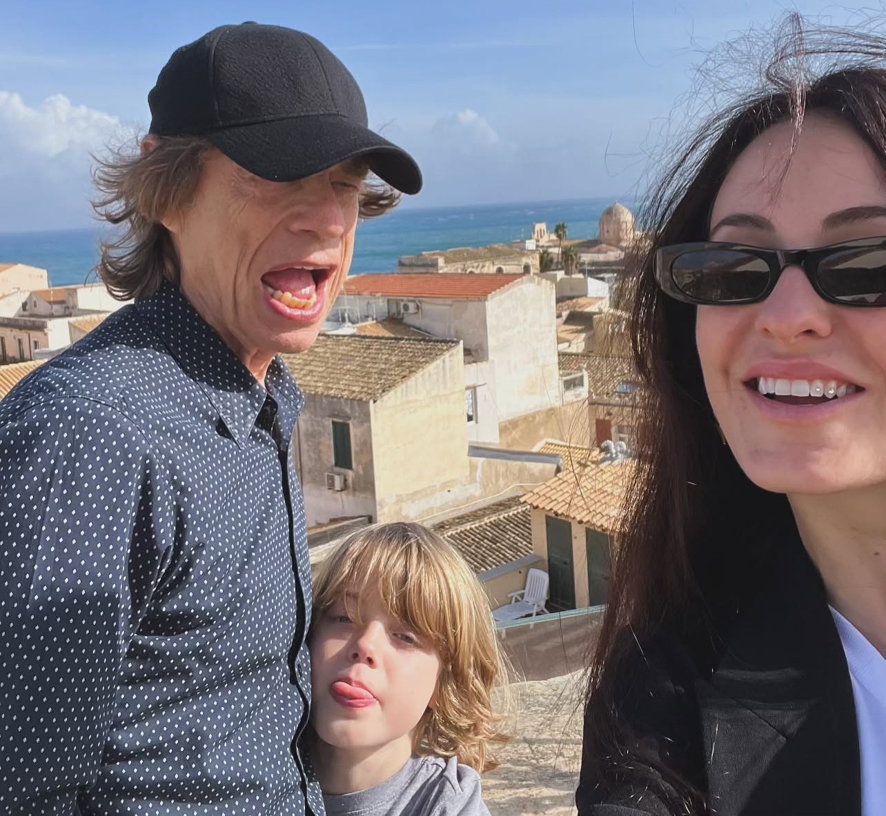 Mick Jagger, Melanie Hamrick, and their son Deveraux posing for a selfie with an ocean and village in the background.