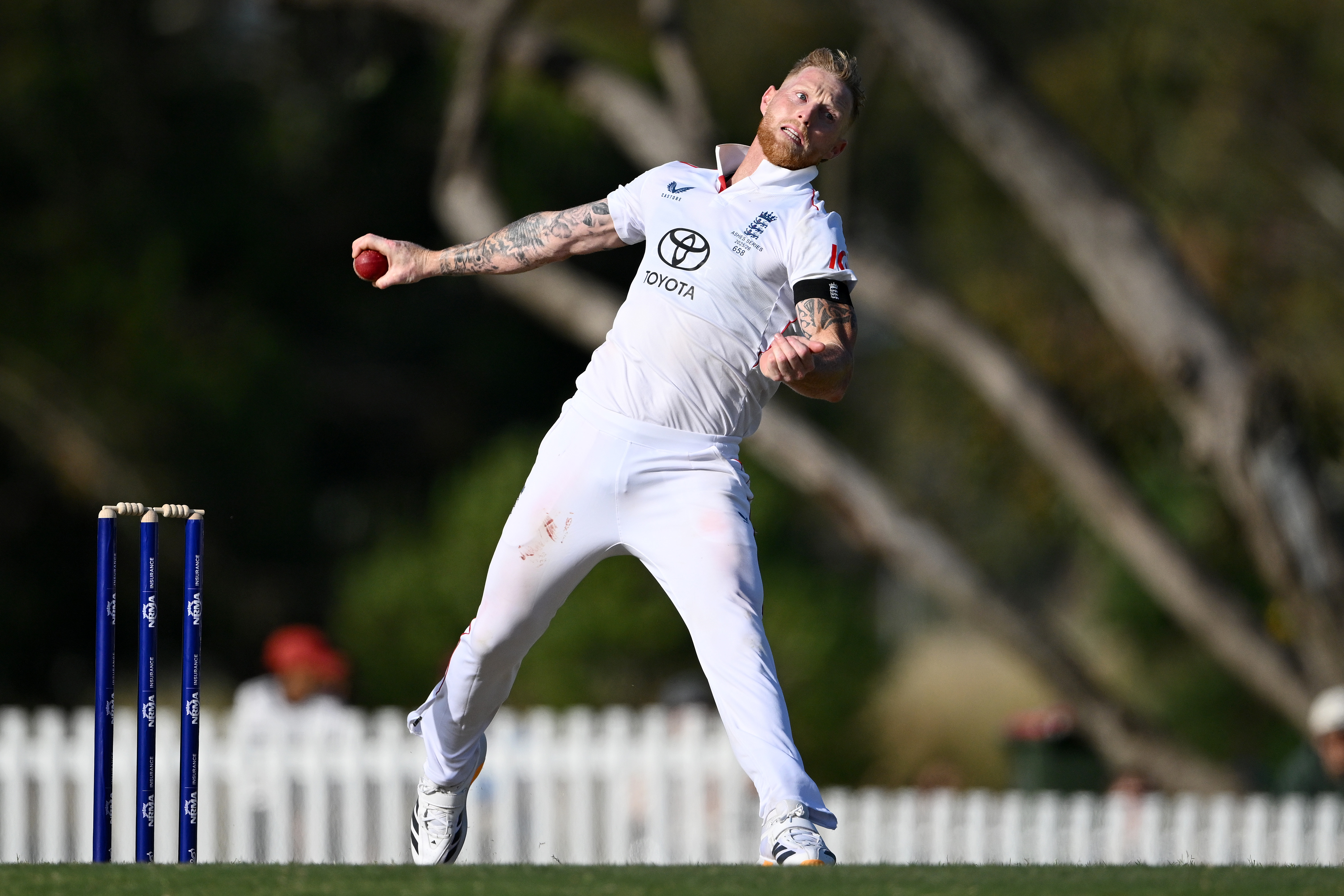 England captain Ben Stokes bowls during a practice match, wearing a white uniform with red dirt marks on his thigh.