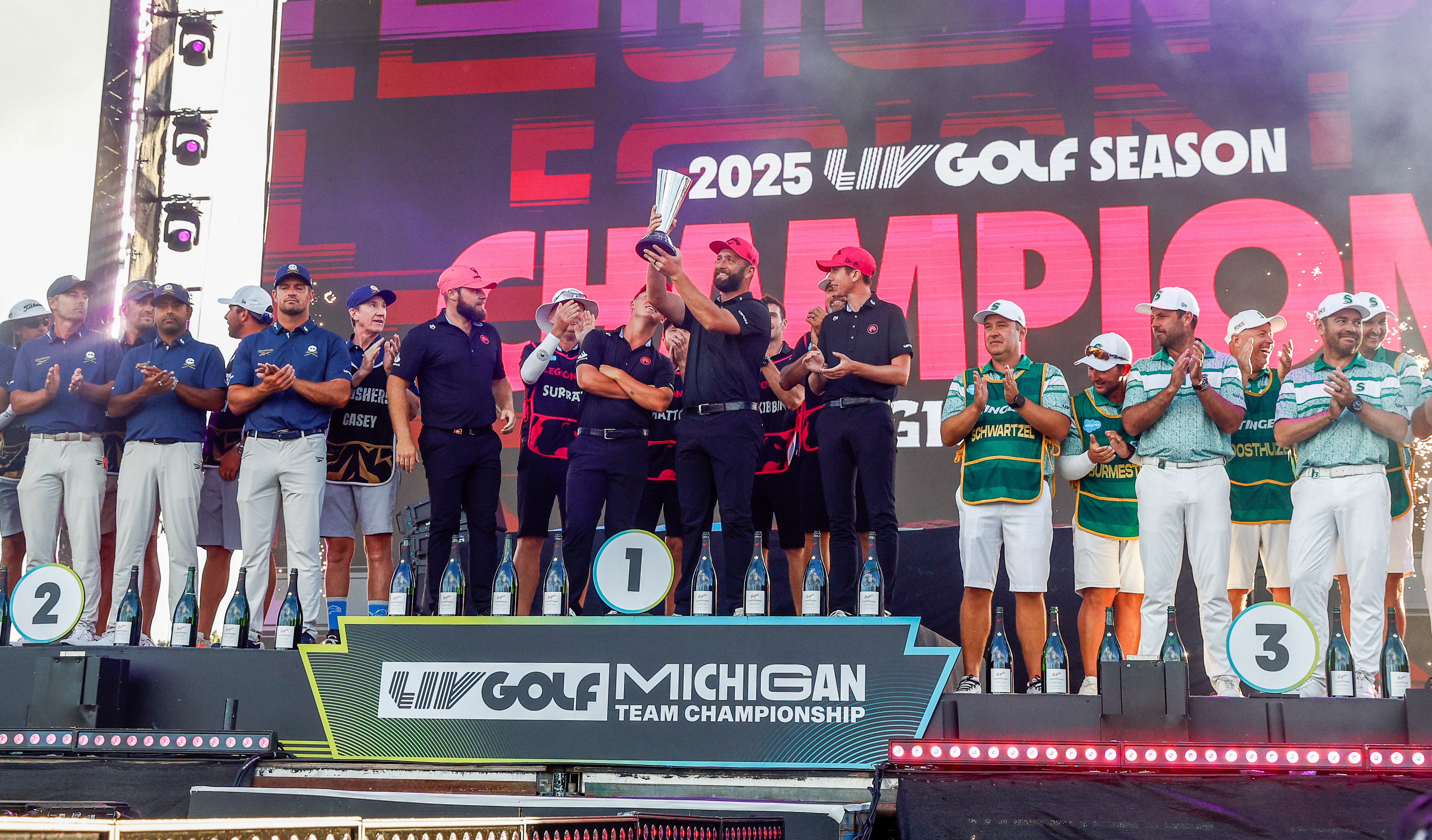 Jon Rahm and his Legion XIII teammates celebrate winning the LIV Golf Championship, raising the trophy on a podium with two other teams.