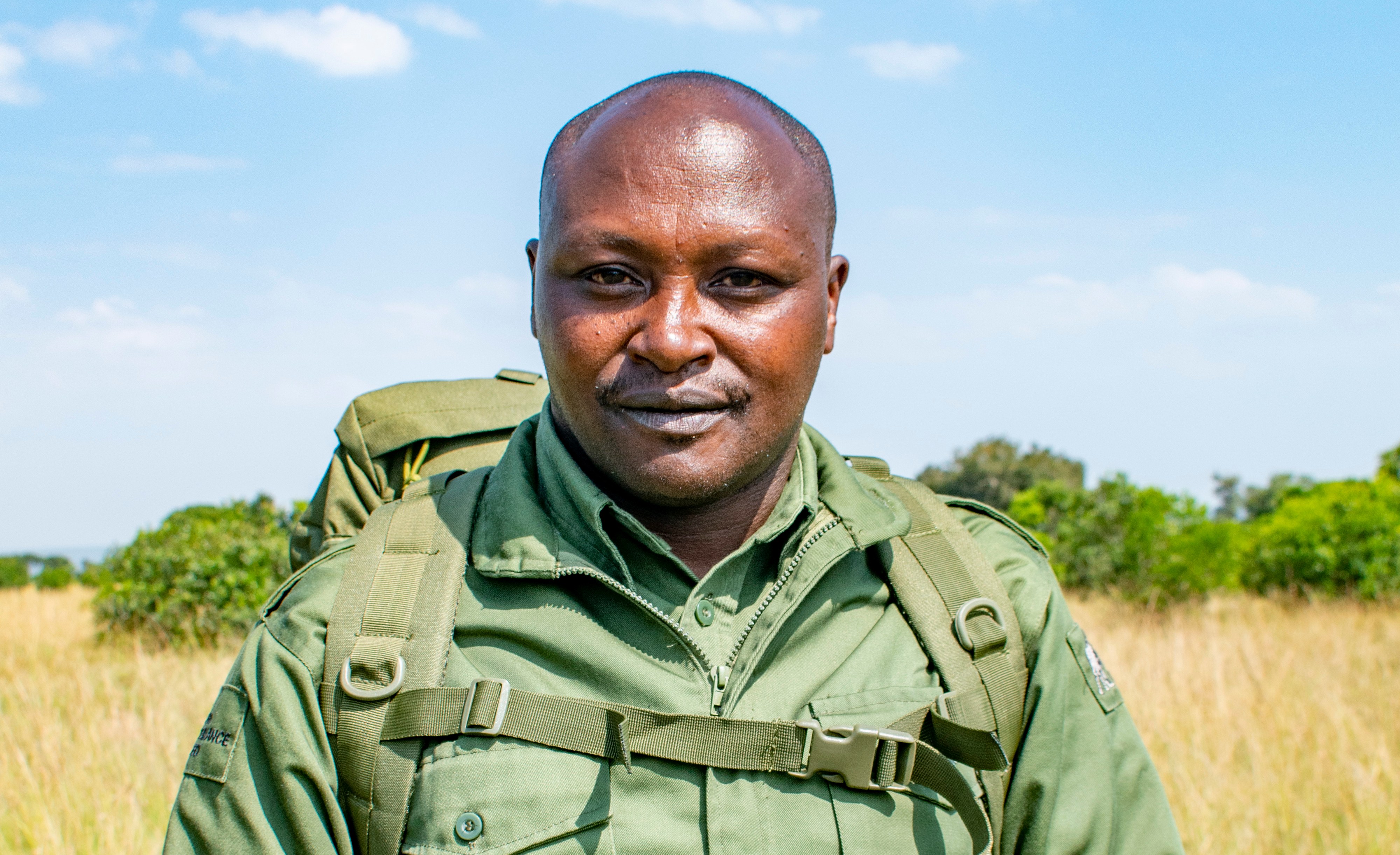 Laban Mwangi, a Kenyan ranger, dressed in an olive green uniform with a backpack, stands in a grassy field.