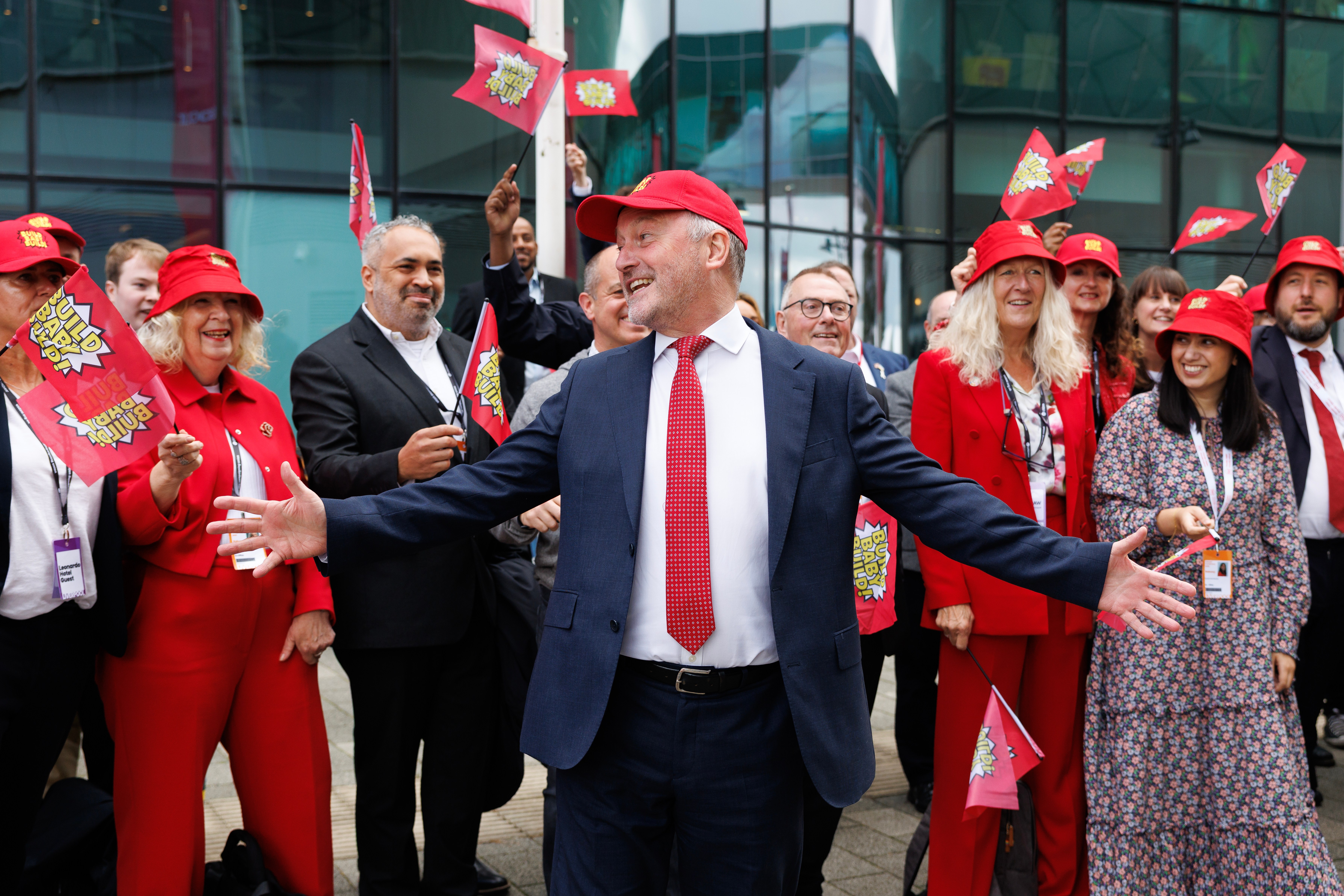 Steve Reed, Secretary of State for Housing, Communities and Local Government, smiles with arms outstretched among supporters holding red flags and wearing red hats.