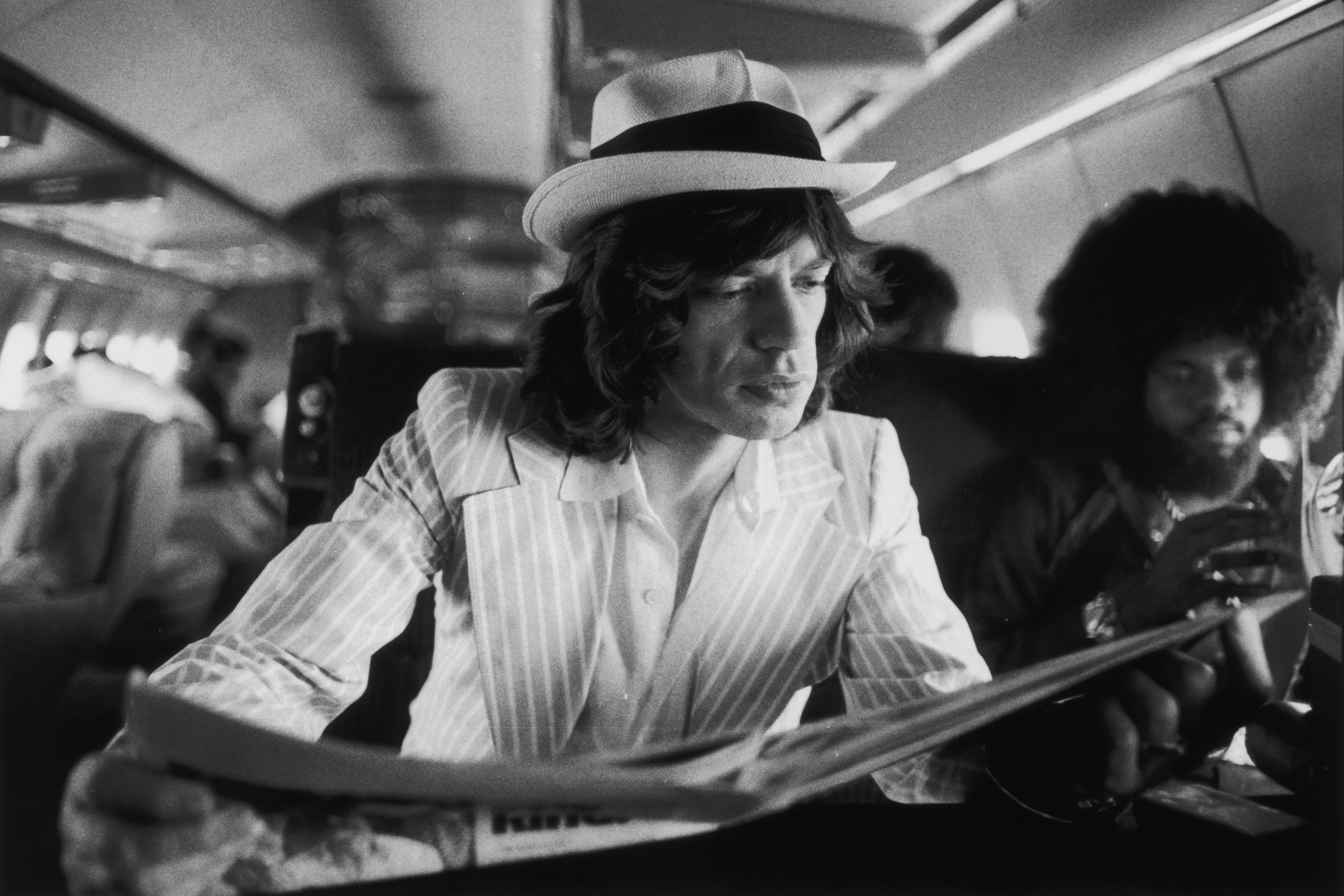 Mick Jagger reading a newspaper while sitting on a private jet with Billy Preston.
