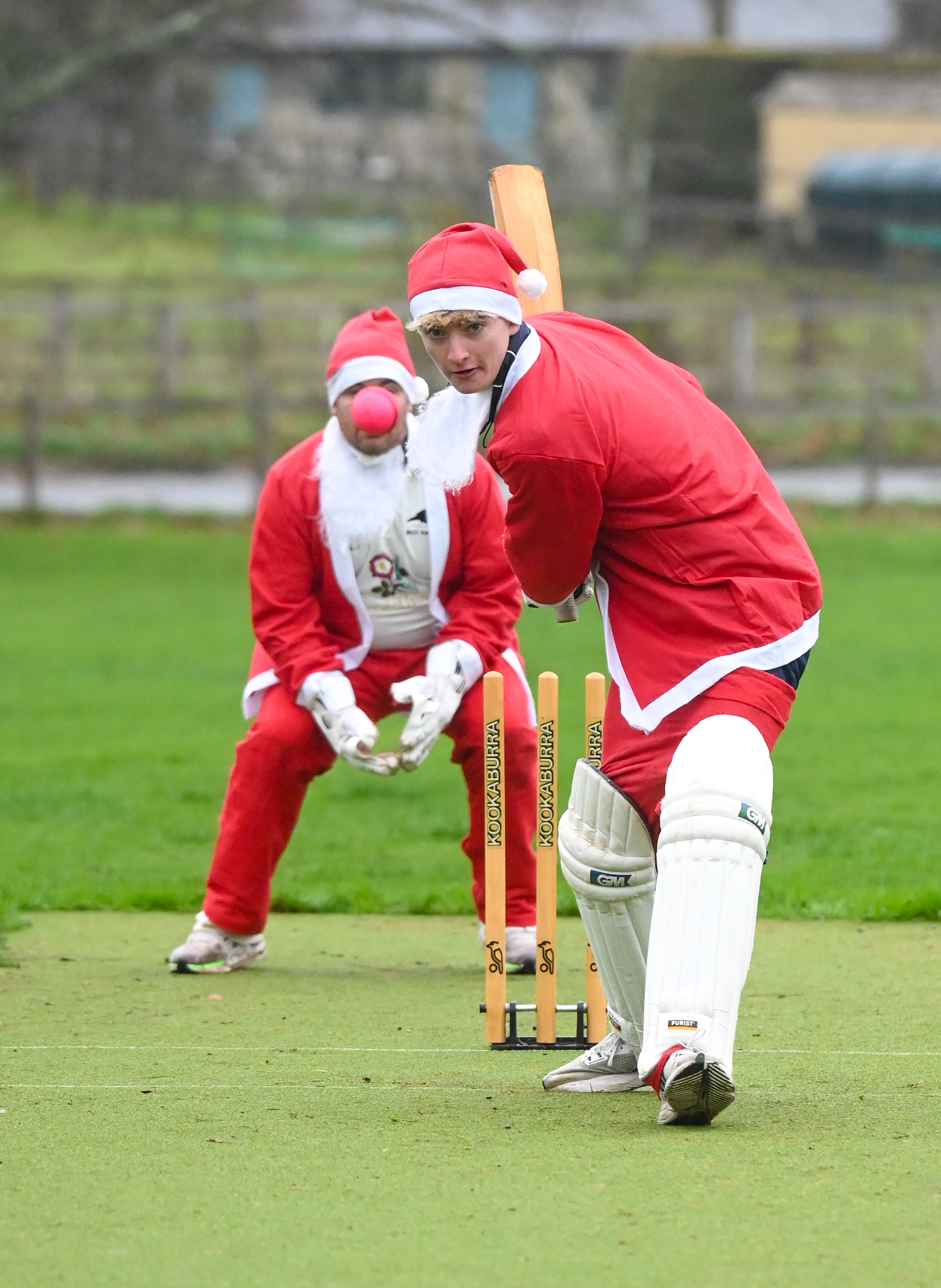 Two men dressed as Santa Claus playing cricket, one batting and one fielding, on a green field.
