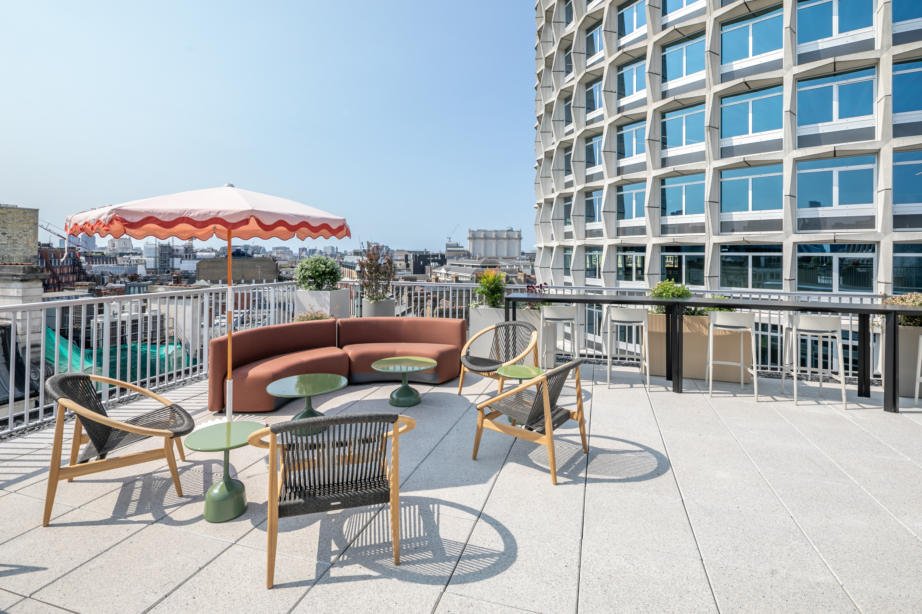 Rooftop terrace with seating area, a large curved brown sofa, green side tables, a patterned parasol, and a geometric-patterned building in the background.