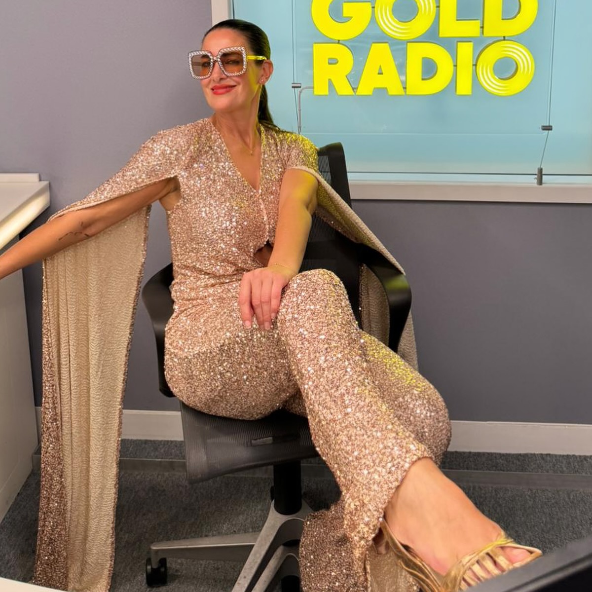 Kirsty Gallacher sitting in a chair, wearing a sparkling gold jumpsuit, gold sandals, and bedazzled sunglasses.