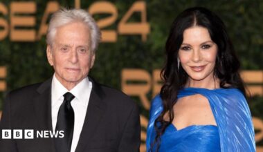 Michael Douglas and Catherine Zeta-Jones are standing next to each other facing towards the camera smiling. Catherine is wearing a blue dress and Michael is wearing a black suite with a white shirt and black tie.