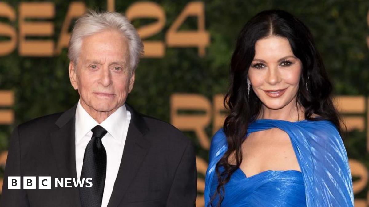 Michael Douglas and Catherine Zeta-Jones are standing next to each other facing towards the camera smiling. Catherine is wearing a blue dress and Michael is wearing a black suite with a white shirt and black tie.
