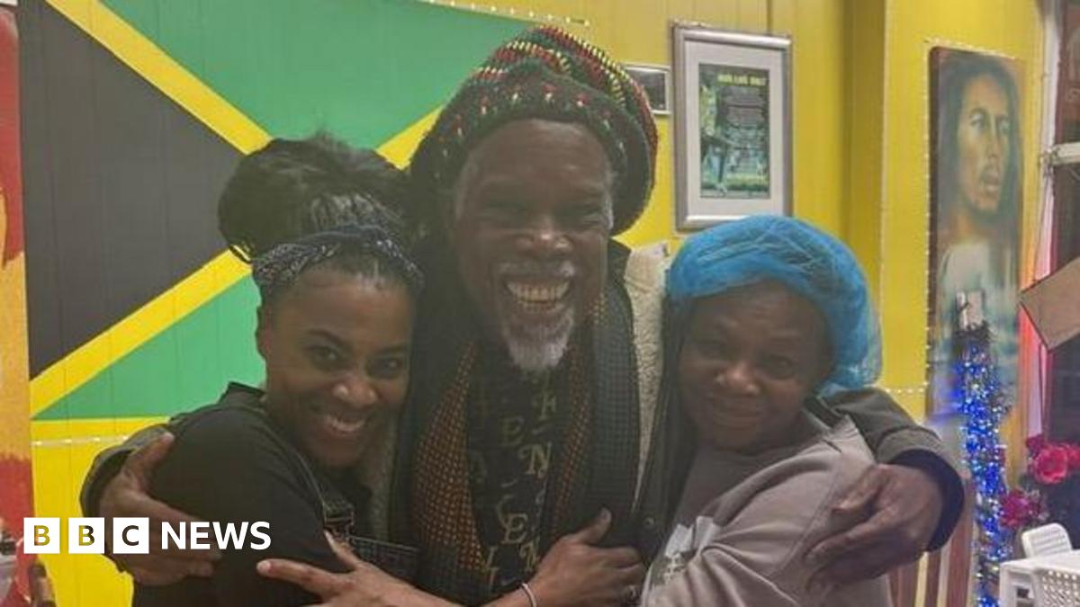 Three people are standing closely together inside a brightly lit room, embracing each other in a group hug. The person in the center is wearing a colorful patterned outfit with a knitted hat featuring red, green, and yellow stripes. The two individuals on either side are dressed casually, one in a dark apron over clothing and the other in a light sweatshirt with a blue head covering. Behind them, a large Jamaican flag is displayed on a yellow wall, along with framed certificates and posters. A white chair and a table are visible to the side, and the floor is tiled in a light pattern.