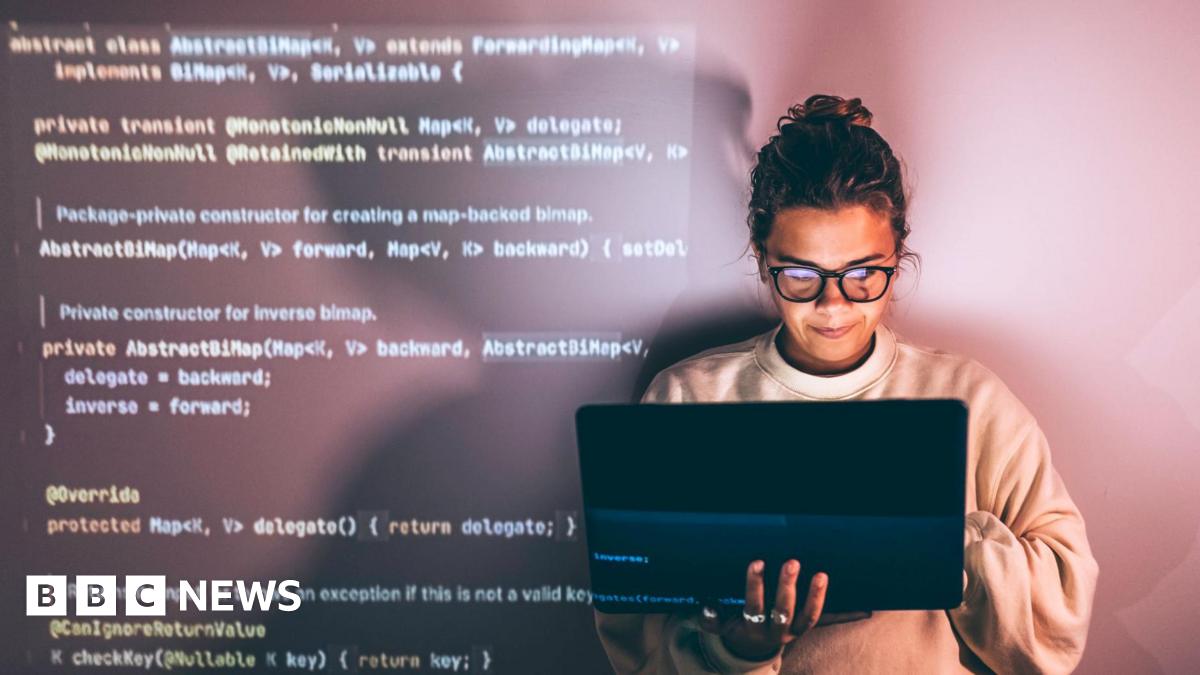 A woman wearing glasses with her hair tied up looks smiling holding a laptop computer in her right hand whilst typing with her left. Programming code appears on the left hand side behind her as a back drop.
