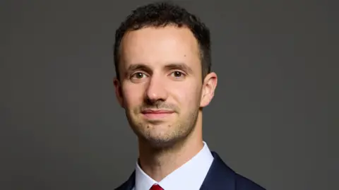 UK Parliament Chris Hinchliff wearing a suit with a white shirt and red tie. He is looking directly at the camera and smiling. 